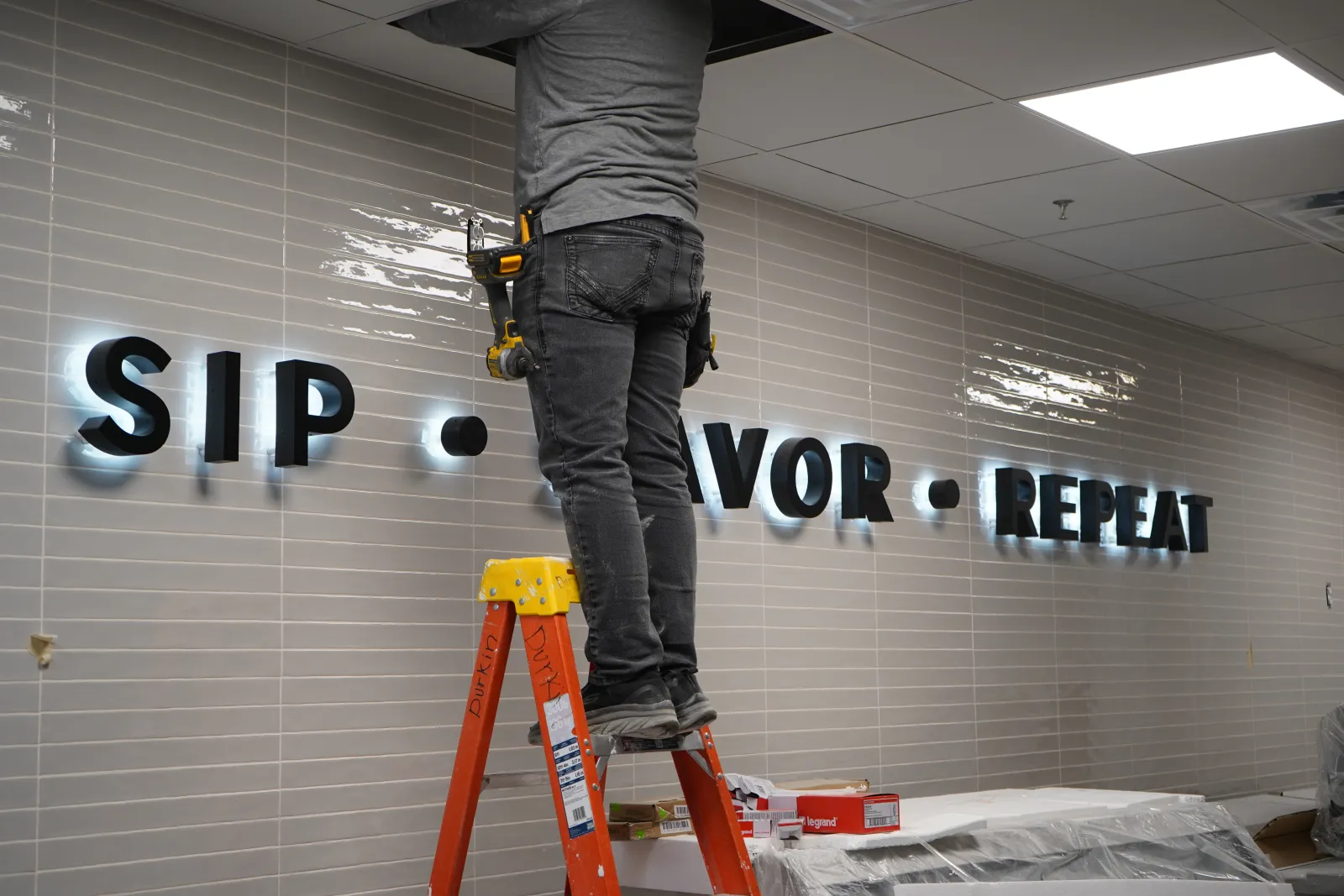Worker on a ladder installing illuminated wall letters reading SIP VOR REPEAT on tiled wall.