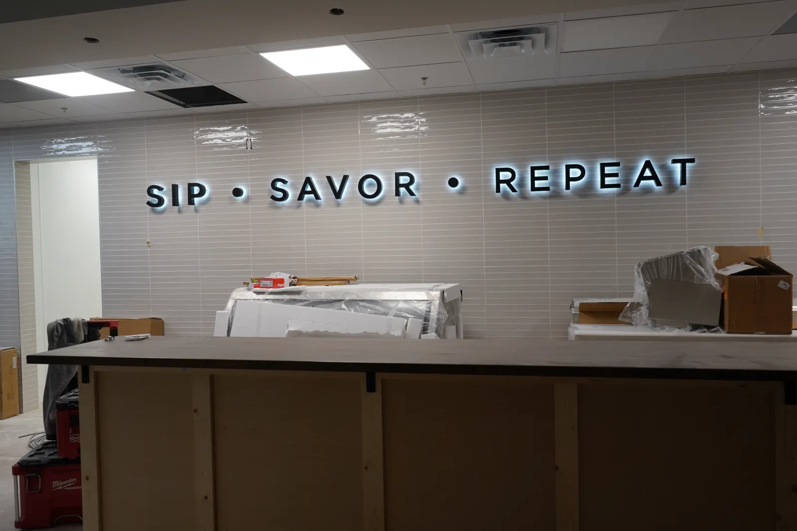 Modern indoor wall with illuminated words SIP • SAVOR • REPEAT above a wooden counter in a new space.