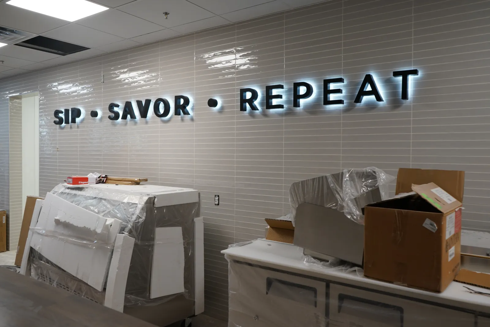Illuminated SIP SAVOR REPEAT sign on tiled wall in room with covered furniture and unpacked boxes.