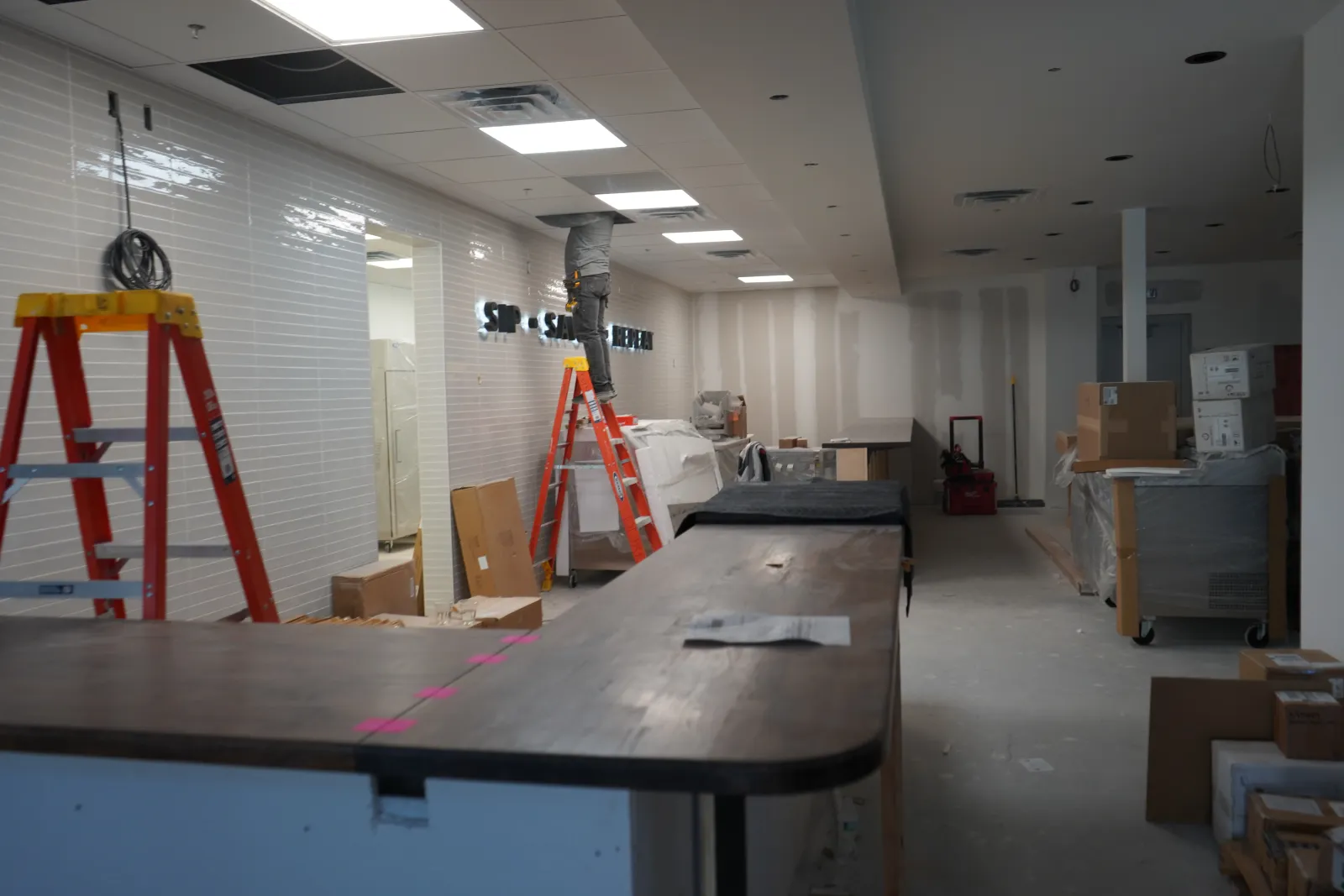 Under-construction interior with ladders, tools, boxes, and a worker installing fixtures in a commercial space.