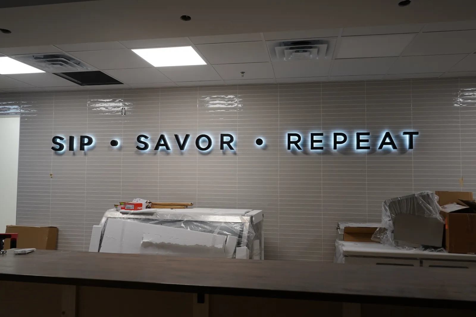 Illuminated wall sign reading SIP SAVOR REPEAT above a cluttered counter with boxes and packing materials.