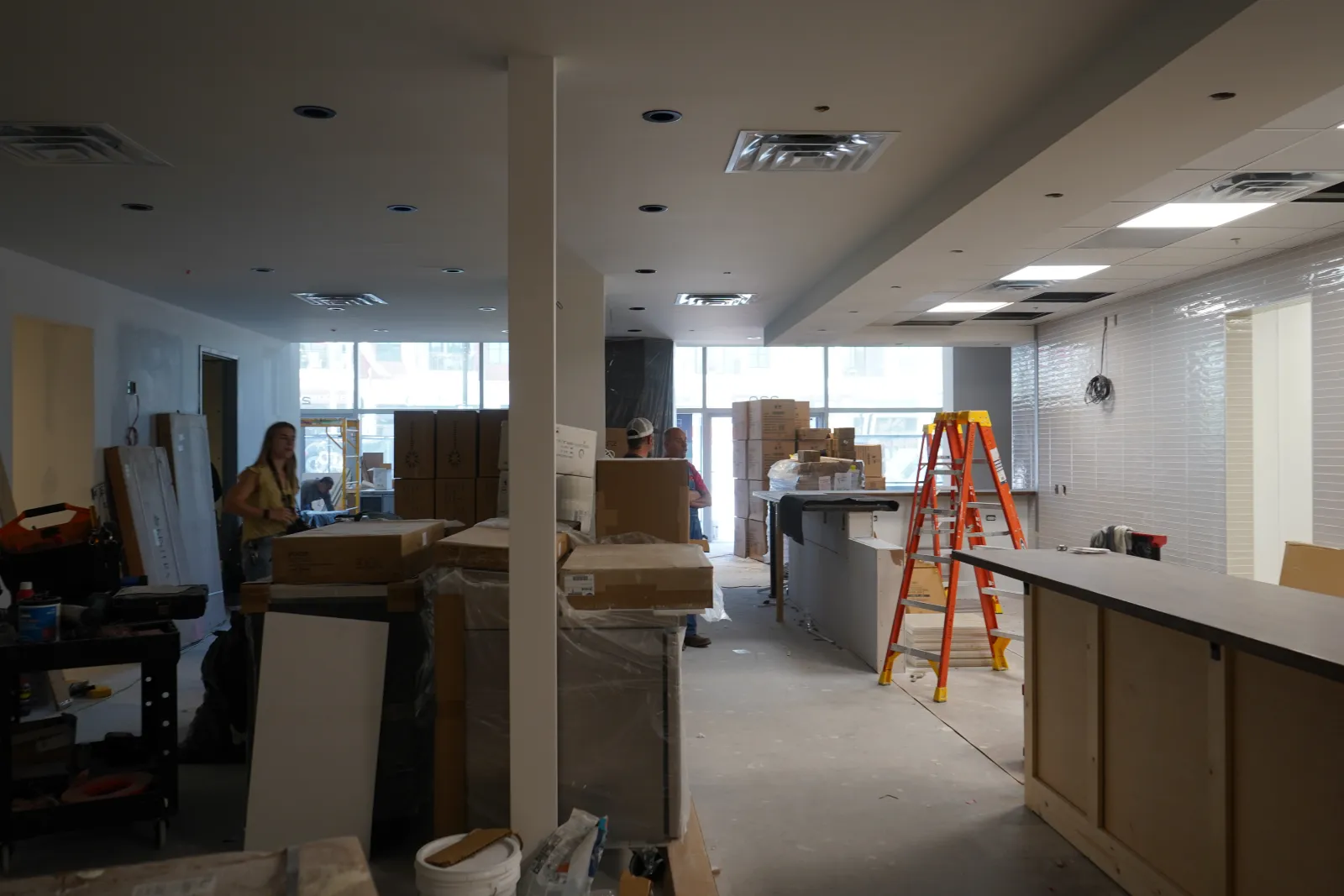 Interior of a commercial space under renovation with stacked boxes, tools, ladders, and workers inside.