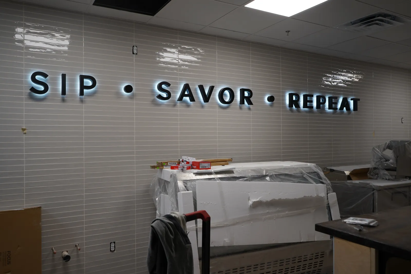 Illuminated wall sign reading SIP • SAVOR • REPEAT in a modern interior under construction with covered furniture.