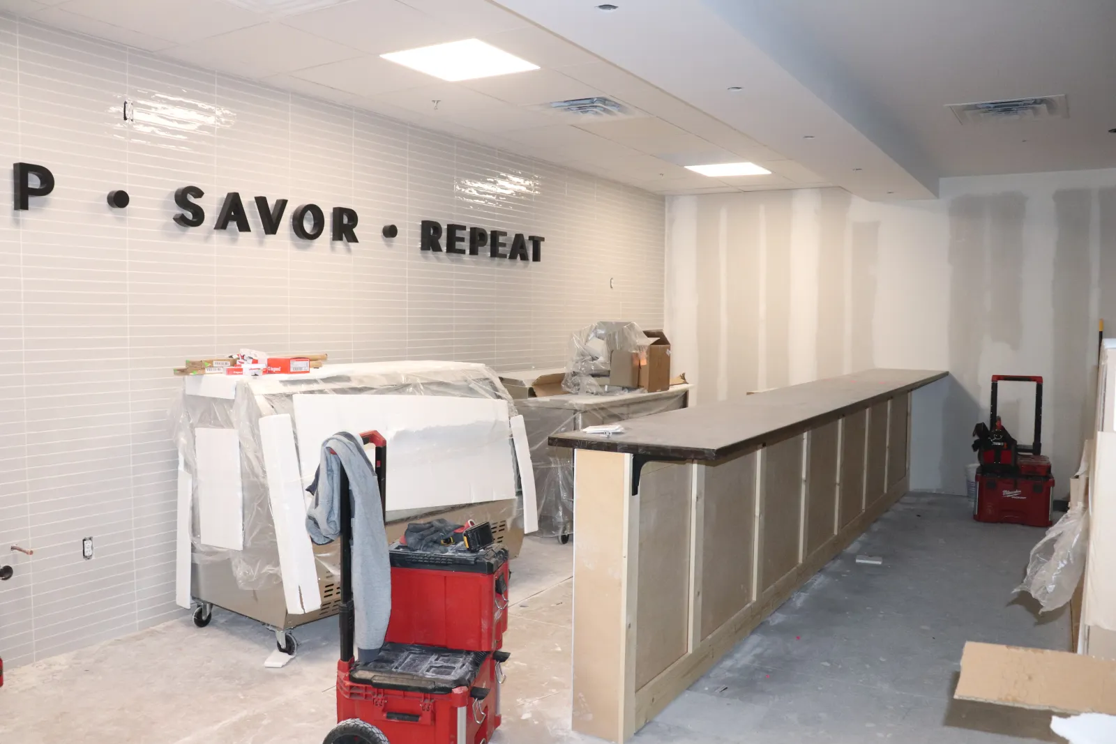 Under-construction restaurant interior with a long counter and 'SAVOR REPEAT' wall signage being installed.