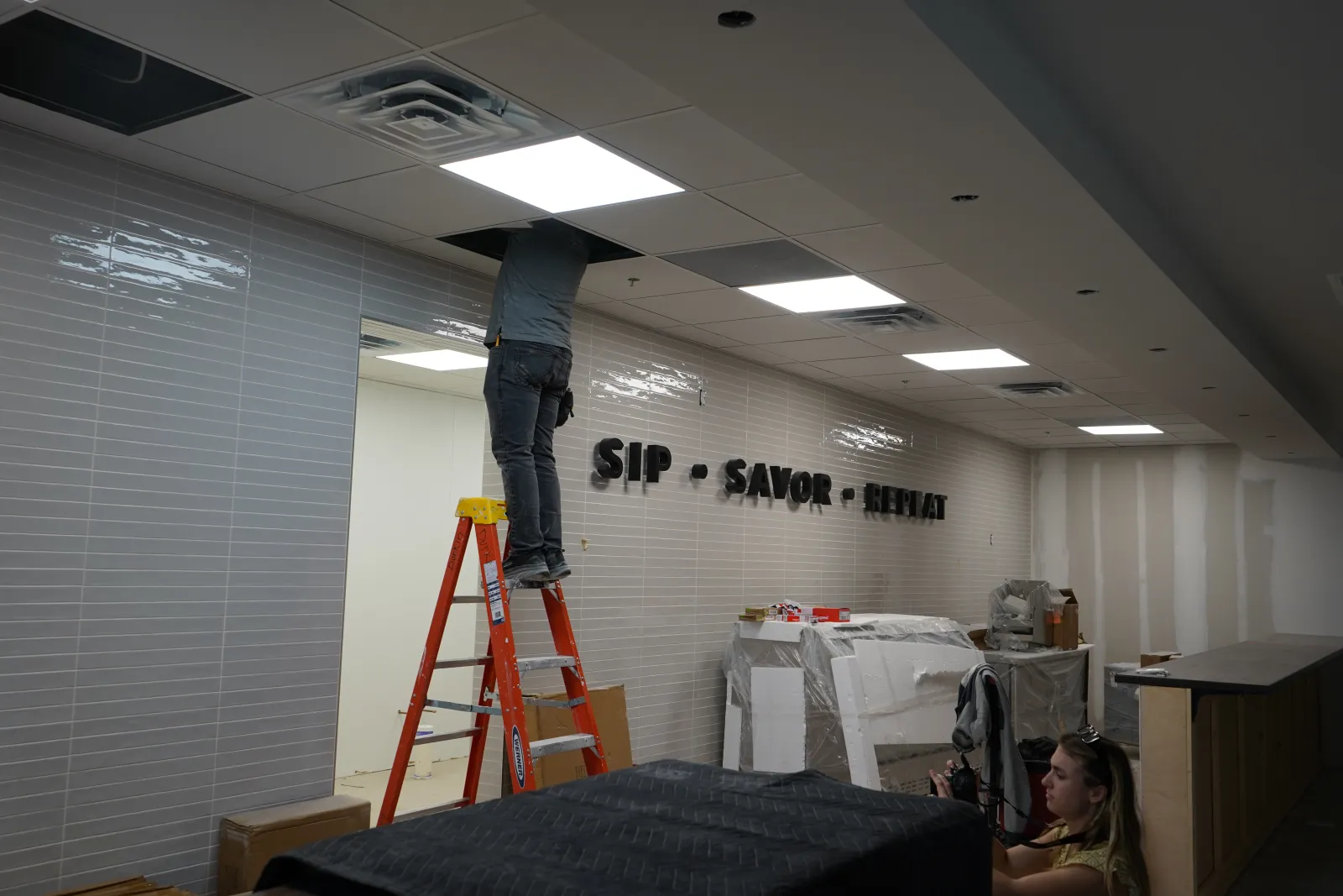 Worker on ladder installing ceiling tiles in a room with tiled walls displaying 'SIP SAVOR RELAX' sign.