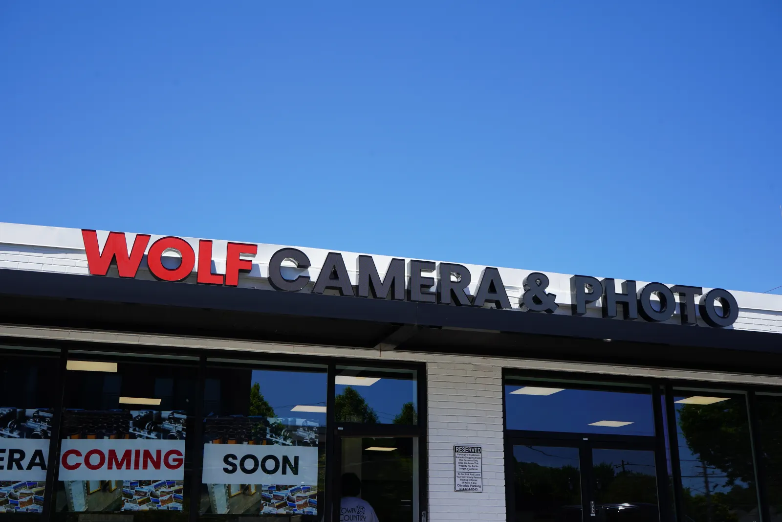 Storefront of Wolf Camera & Photo with a clear blue sky and Coming Soon signs in the windows.