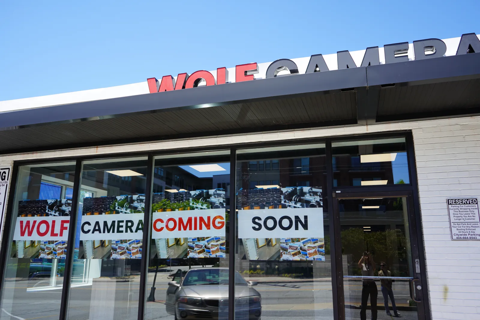 Storefront with signs in windows announcing Wolf Camera coming soon under a clear blue sky.
