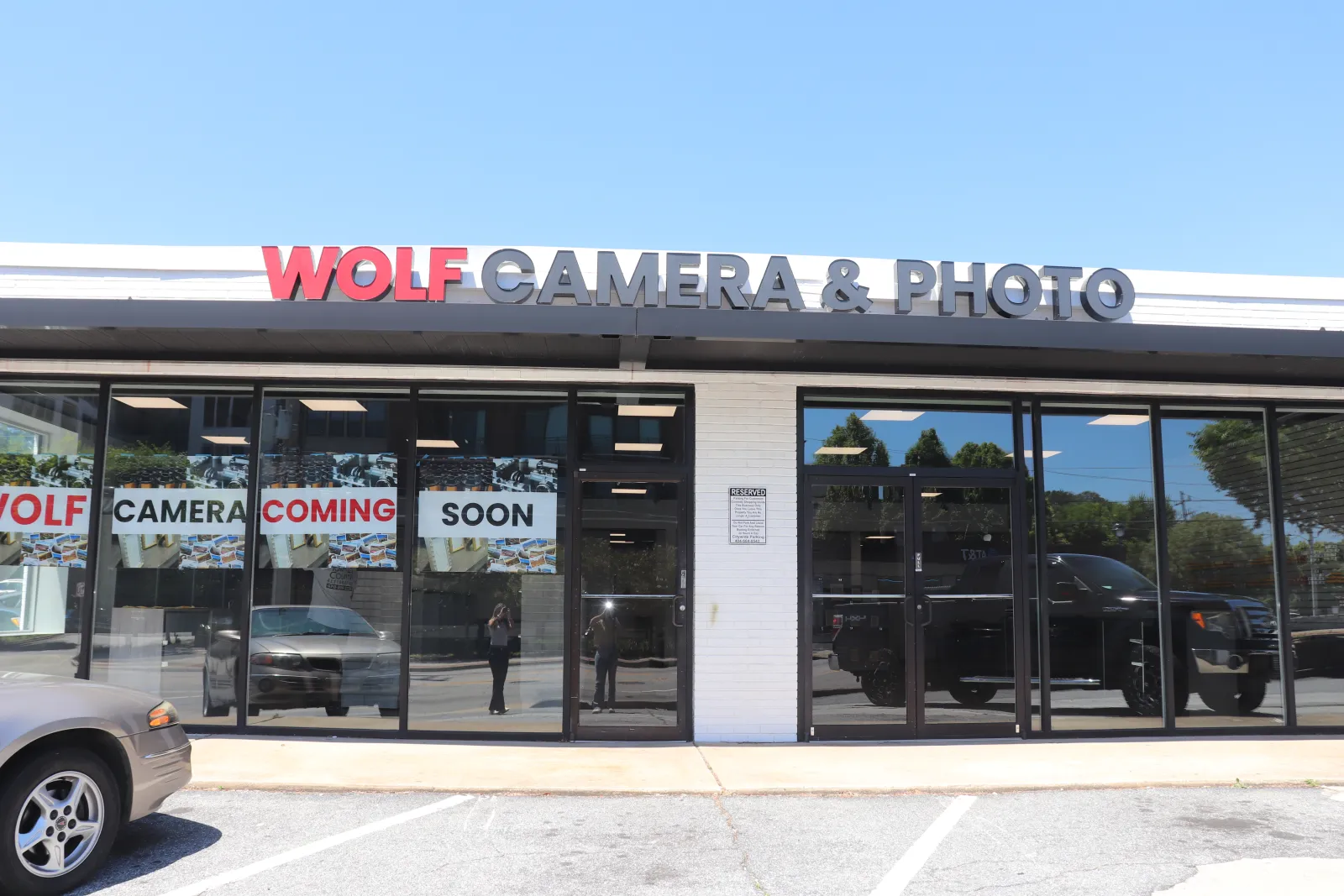Storefront of Wolf Camera & Photo with signs announcing the camera store is coming soon under clear blue sky.