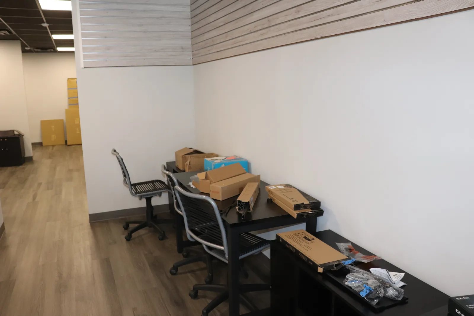 Office space with black tables and chairs cluttered with cardboard boxes and packaging supplies on wooden floor.
