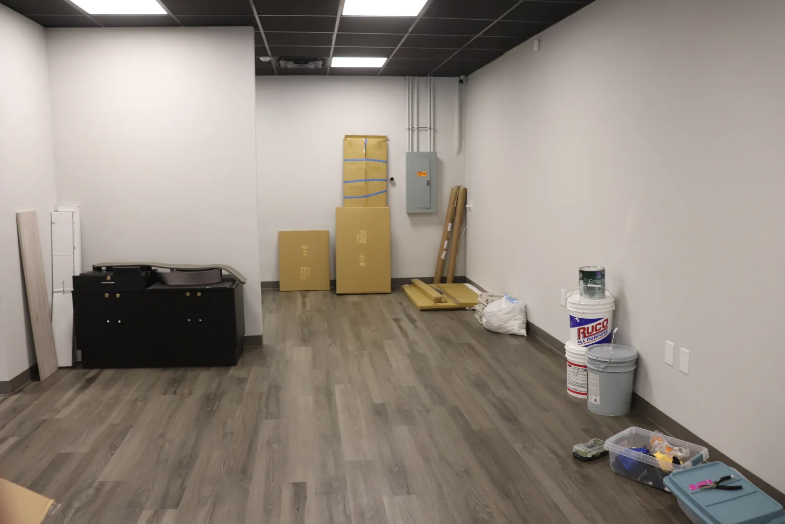 Empty room with gray wood flooring, white walls, black ceiling tiles, and scattered moving boxes and paint supplies.