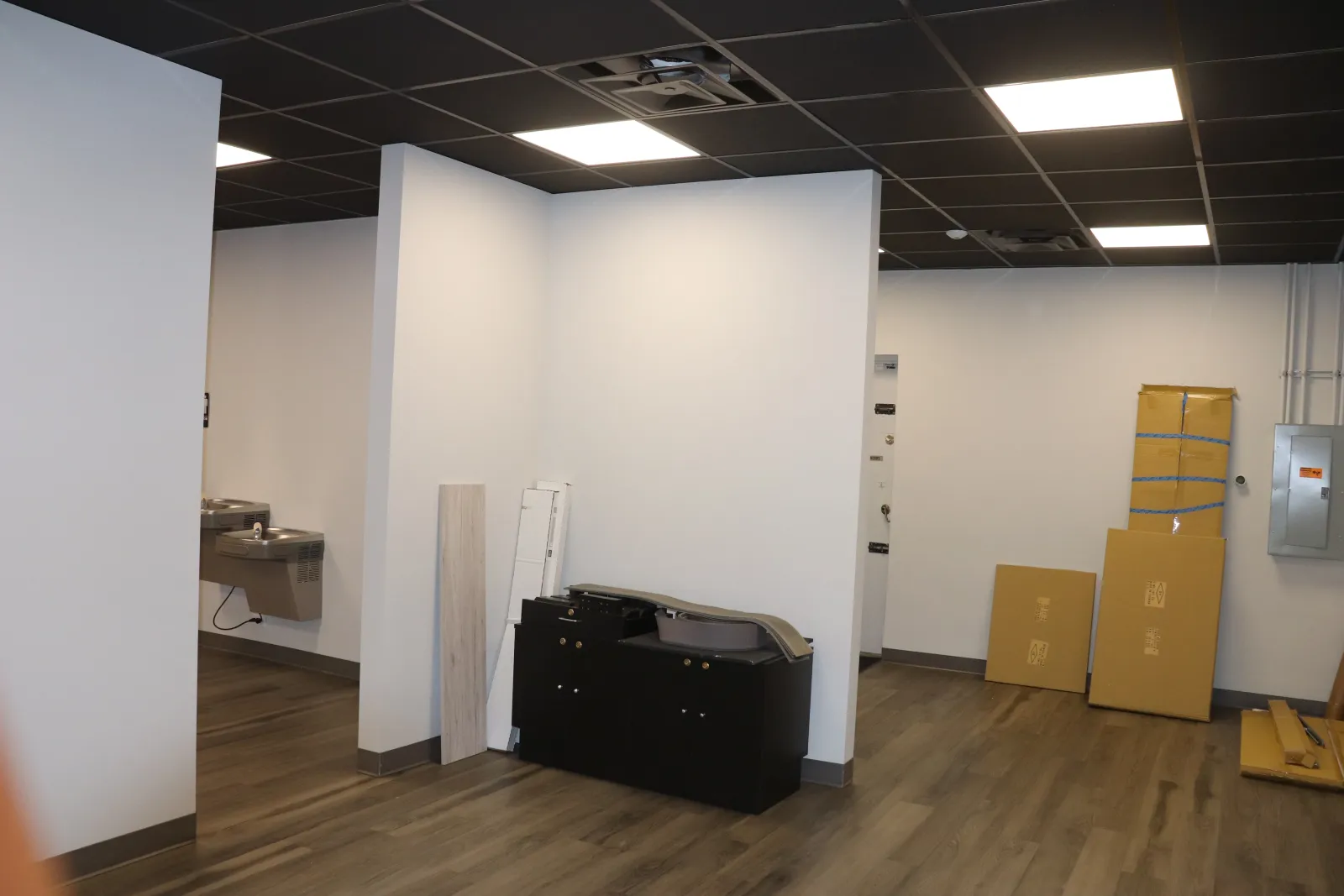 Empty office space with wood flooring, white walls, ceiling lights, drinking fountains, and packed boxes near electrical panels.