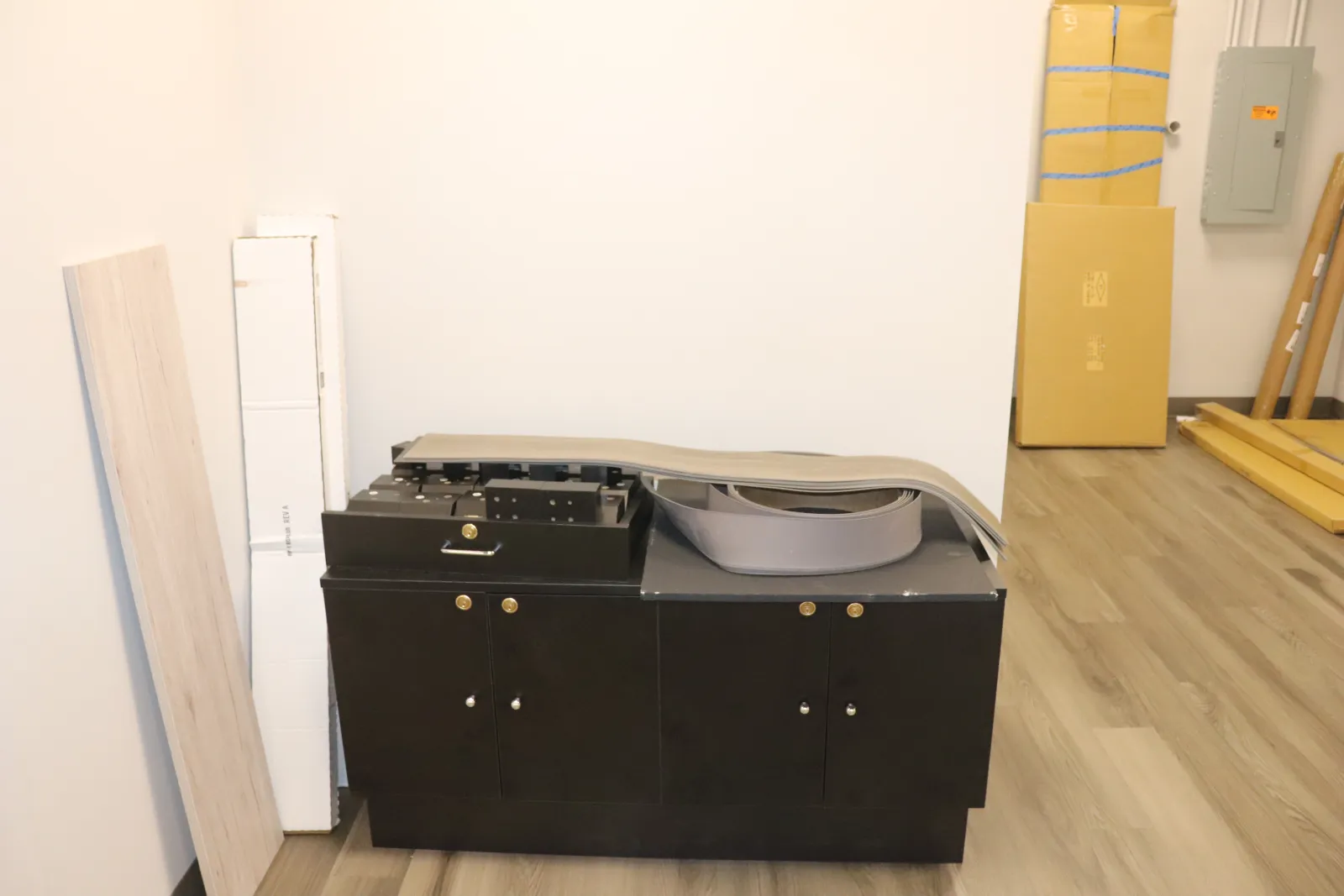 Black printer or copier machine in a room with wooden floor and white walls, surrounded by stacked boxes and panels