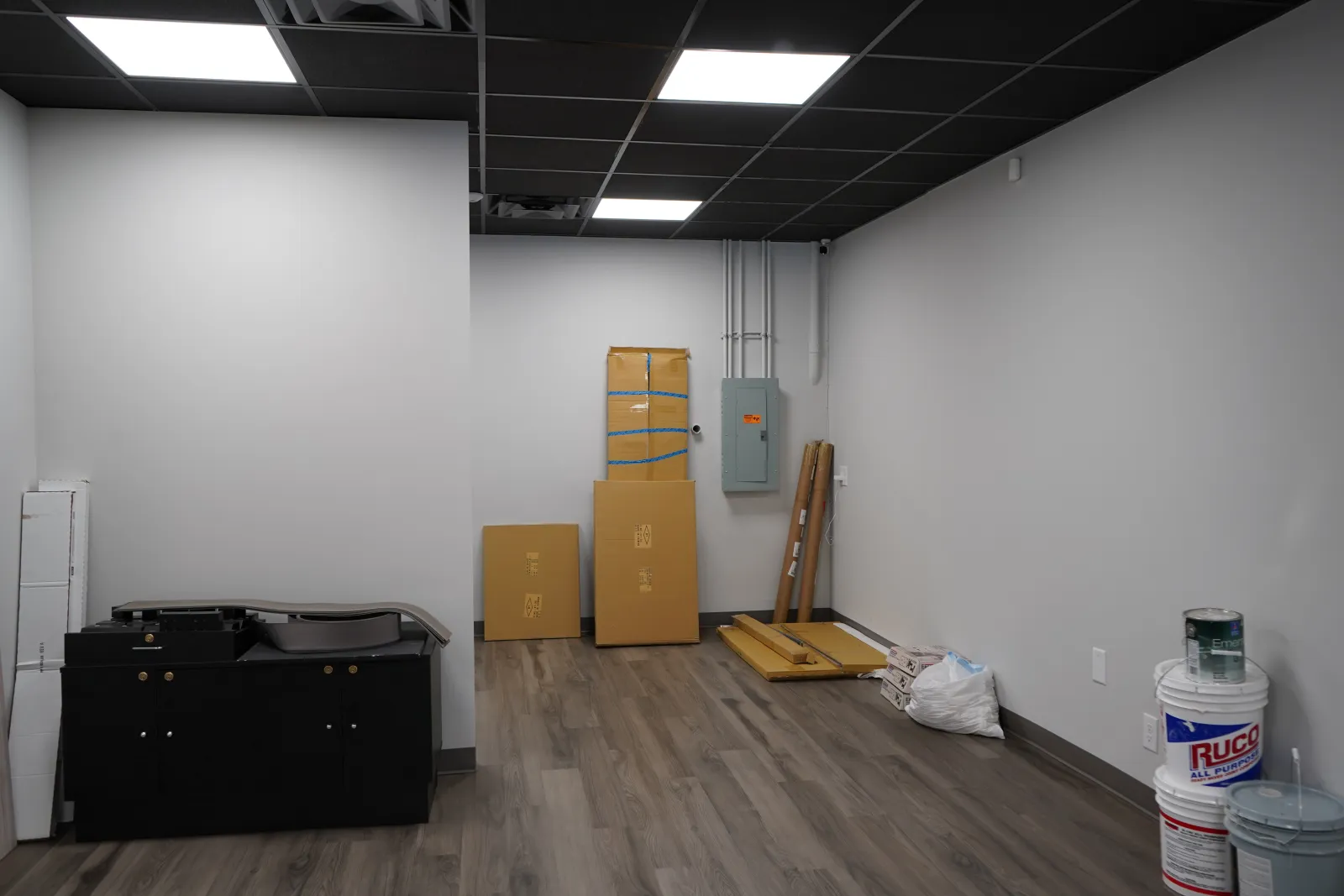 Empty room with light wood flooring, gray walls, black ceiling tiles, cardboard boxes, and paint supplies on the floor.