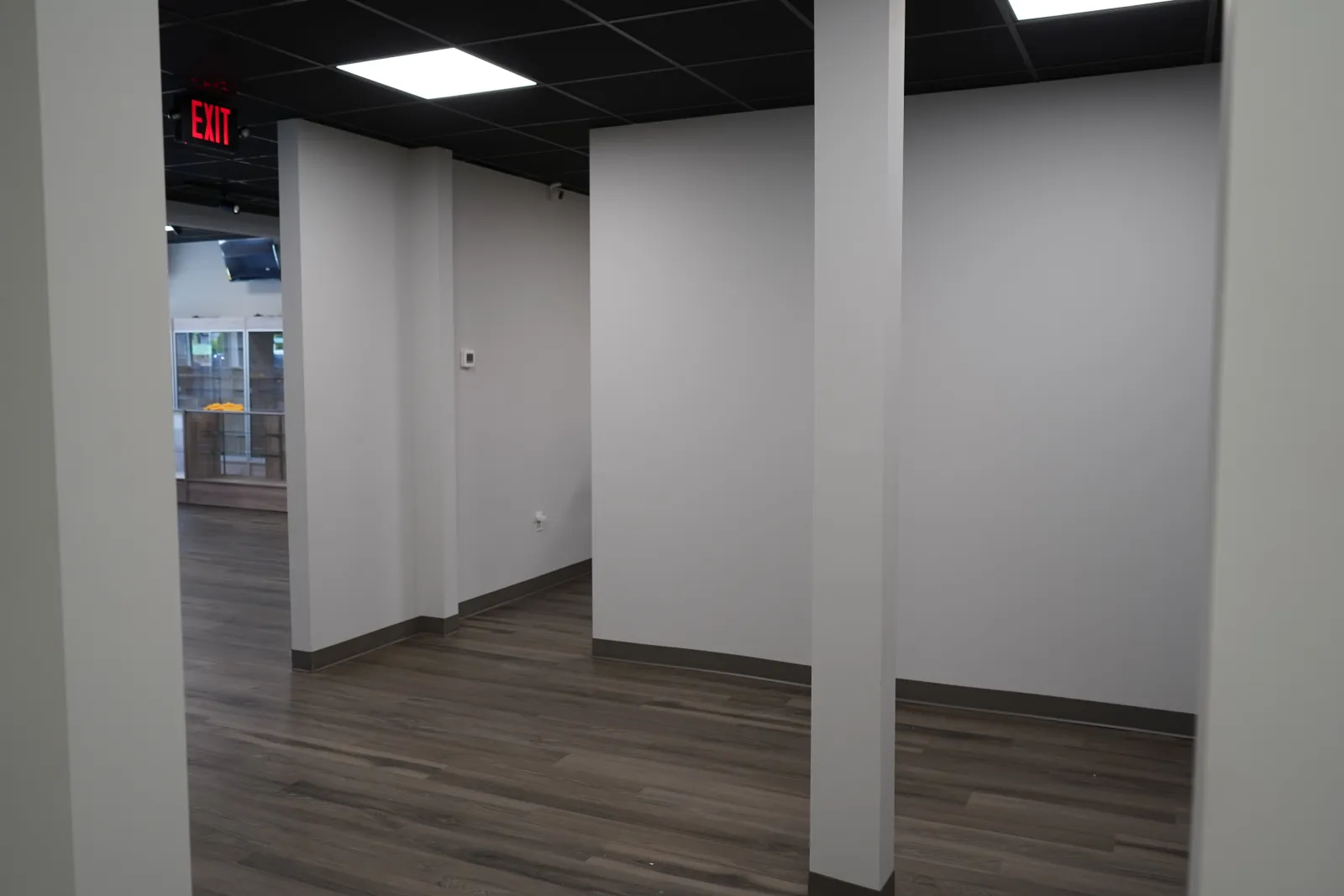 Empty office space with gray walls, wood flooring, and black tiled ceiling with exit sign.