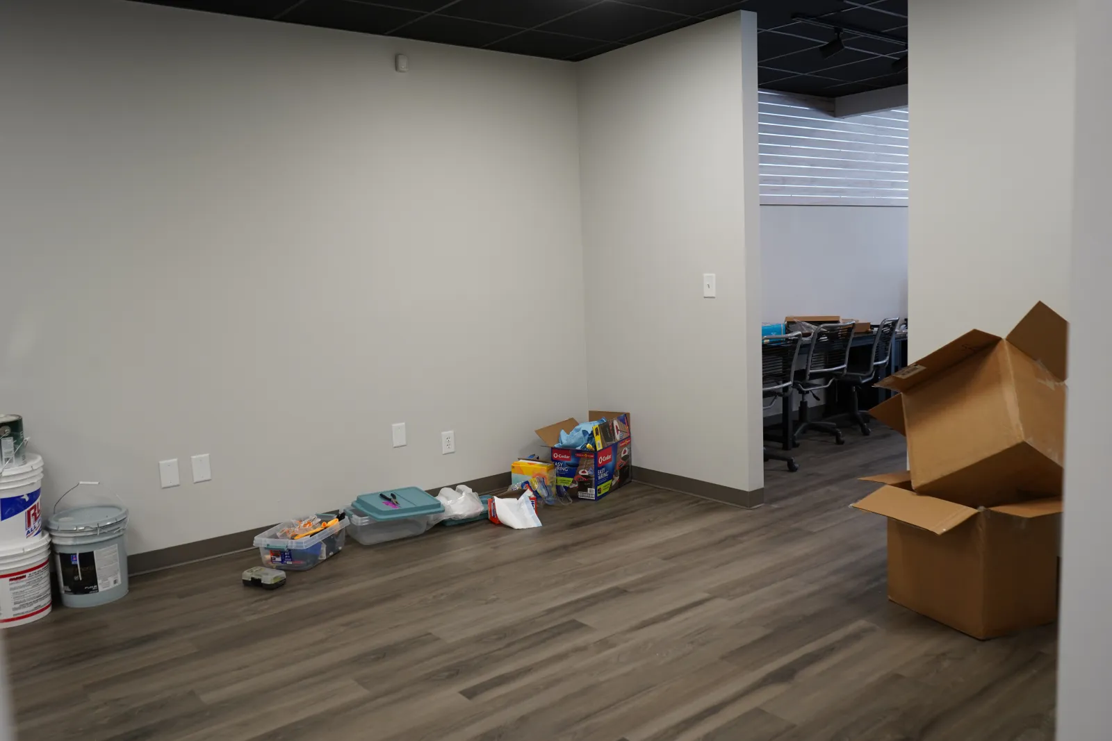 Empty office corner with unpacked boxes, paint buckets, and plastic containers on wooden floor near white walls.