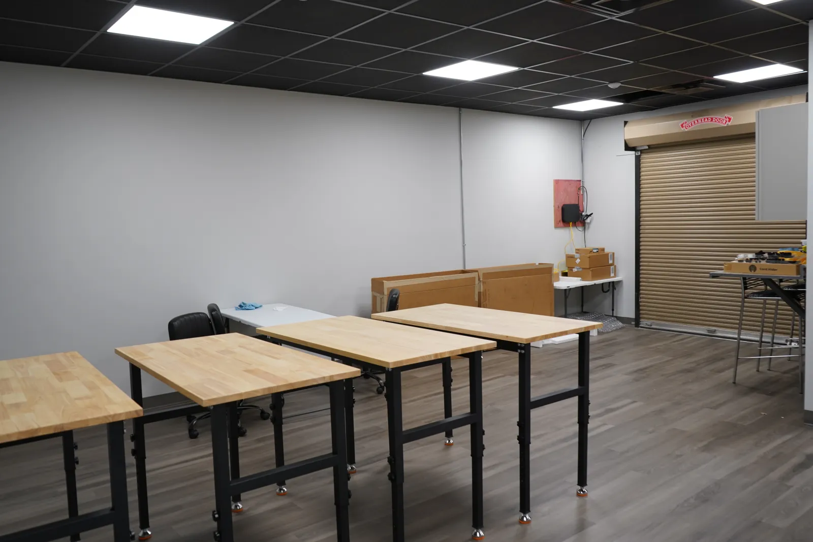 Empty workshop room with wooden tables, black chairs, gray walls, and a roll-up door on wood flooring.