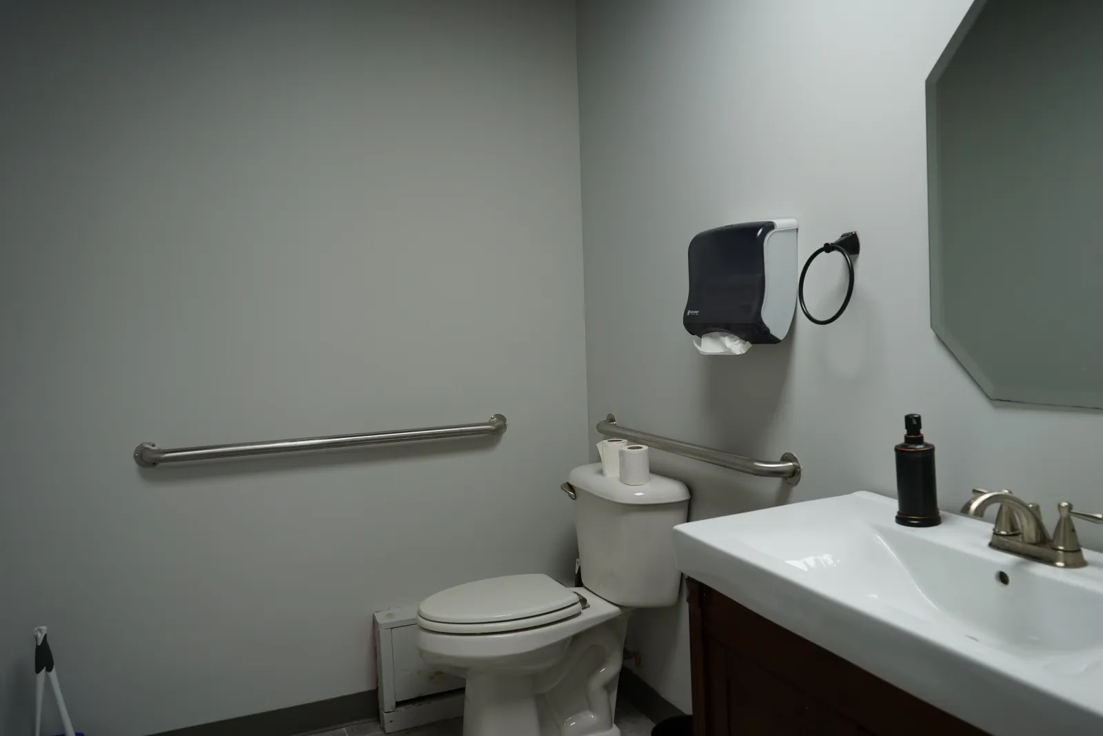 Accessible public restroom with toilet, sink, handrails, paper towel dispenser, and mirror in a clean, simple layout.