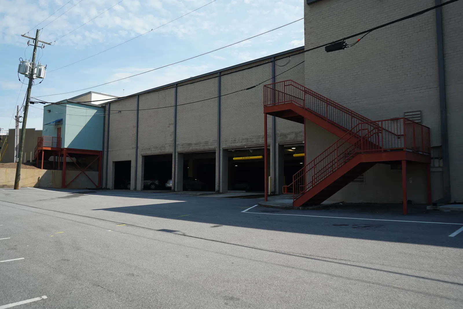 Empty industrial parking lot next to a beige warehouse with red exterior staircases under a blue sky.