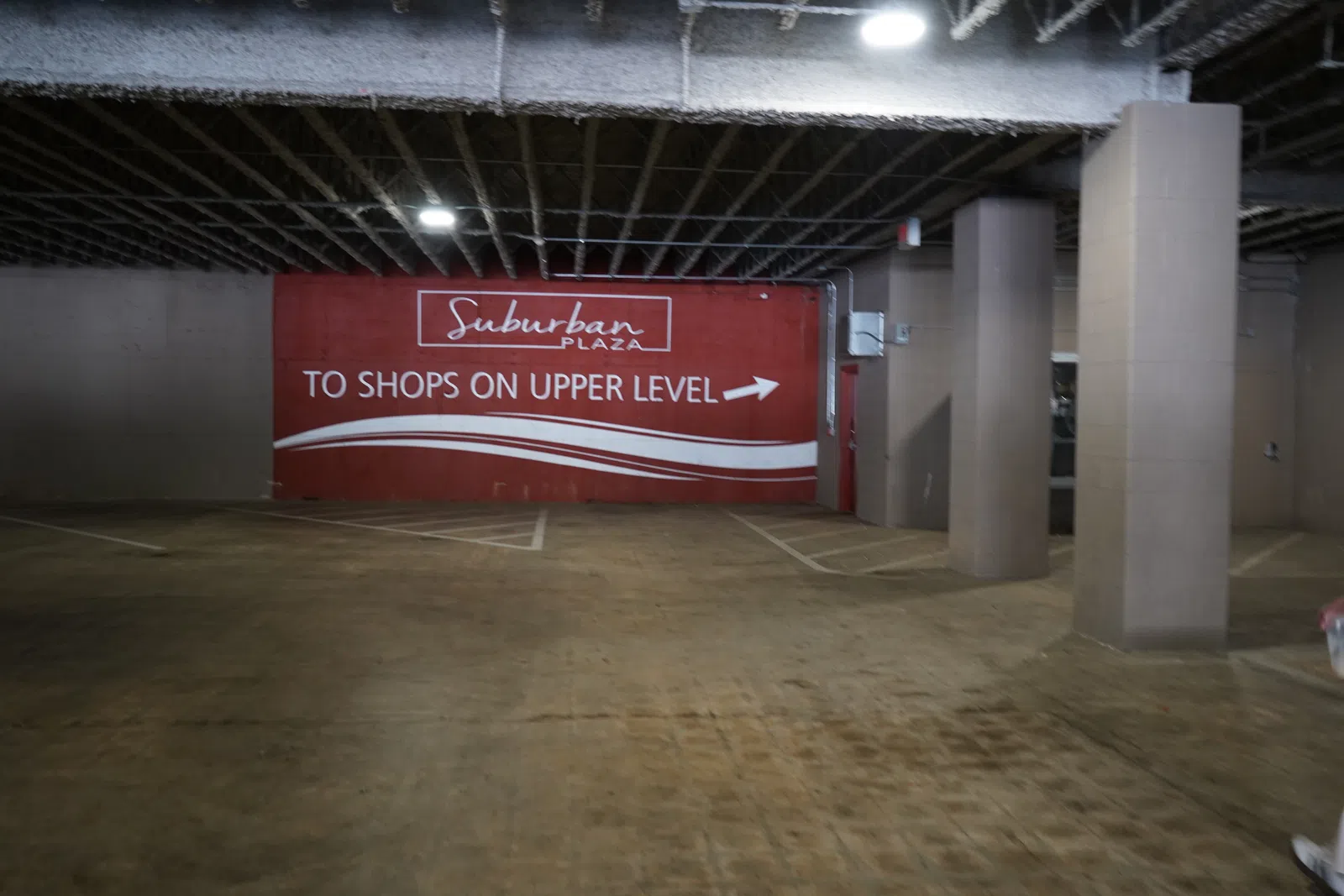 Empty parking garage interior with a red sign directing to Suburban Plaza shops on upper level.