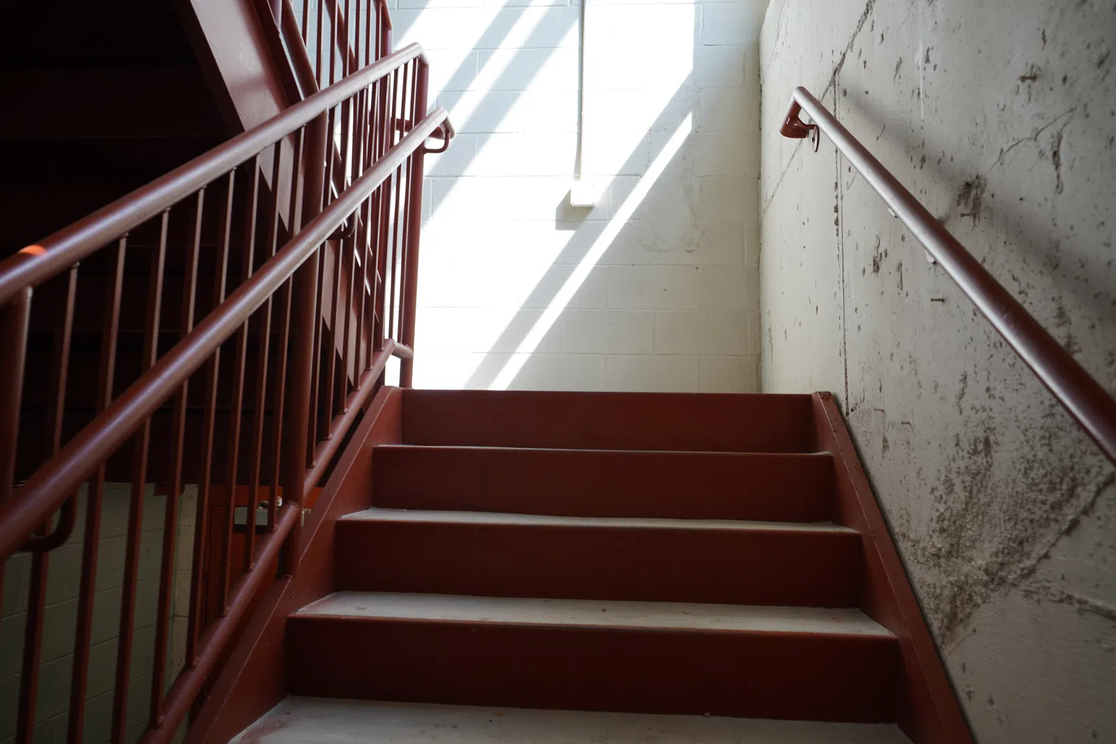 Red metal staircase with handrails leading up to a sunlit white brick wall and window.
