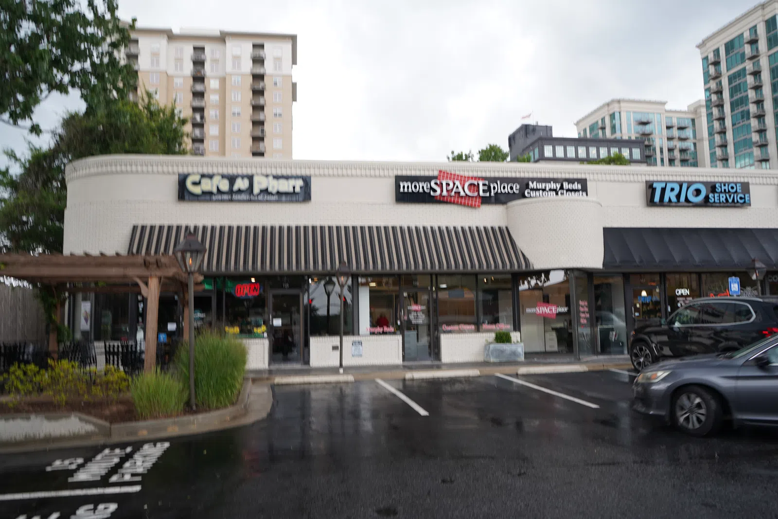 Storefronts of Cafe A Pharr, moreSPACEplace, and TRIO Shoe Service in an urban shopping plaza with parked cars.