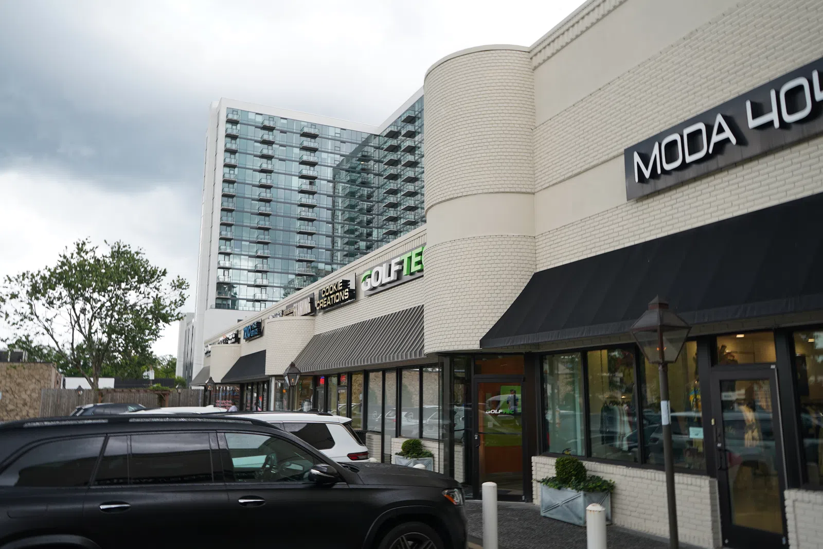 Modern retail strip with Moda House and other stores under cloudy sky and black SUV parked outside.