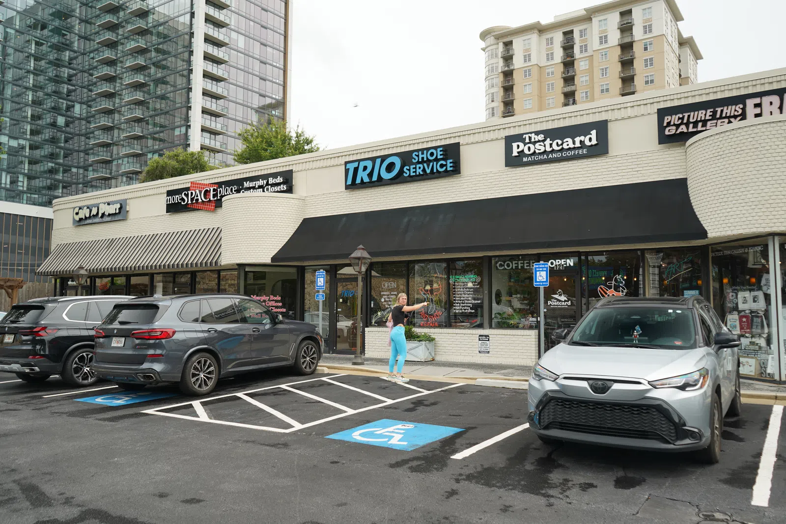 Street view of small retail shops including Trio Shoe Service, The Postcard cafe, and parking with cars and a person in bright pants.