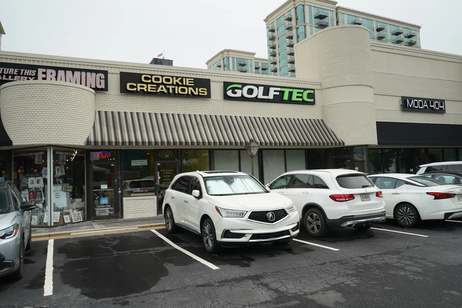 Strip mall storefronts with shops named Cookie Creations, GolfTec, and Moda 404 with cars parked in front on a cloudy day