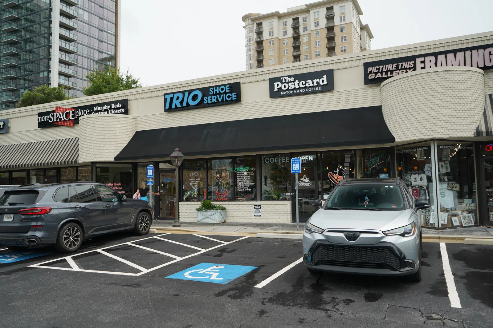 Storefronts of Trio Shoe Service, The Postcard coffee shop, and Picture This Framing with parked cars and accessible parking spaces.