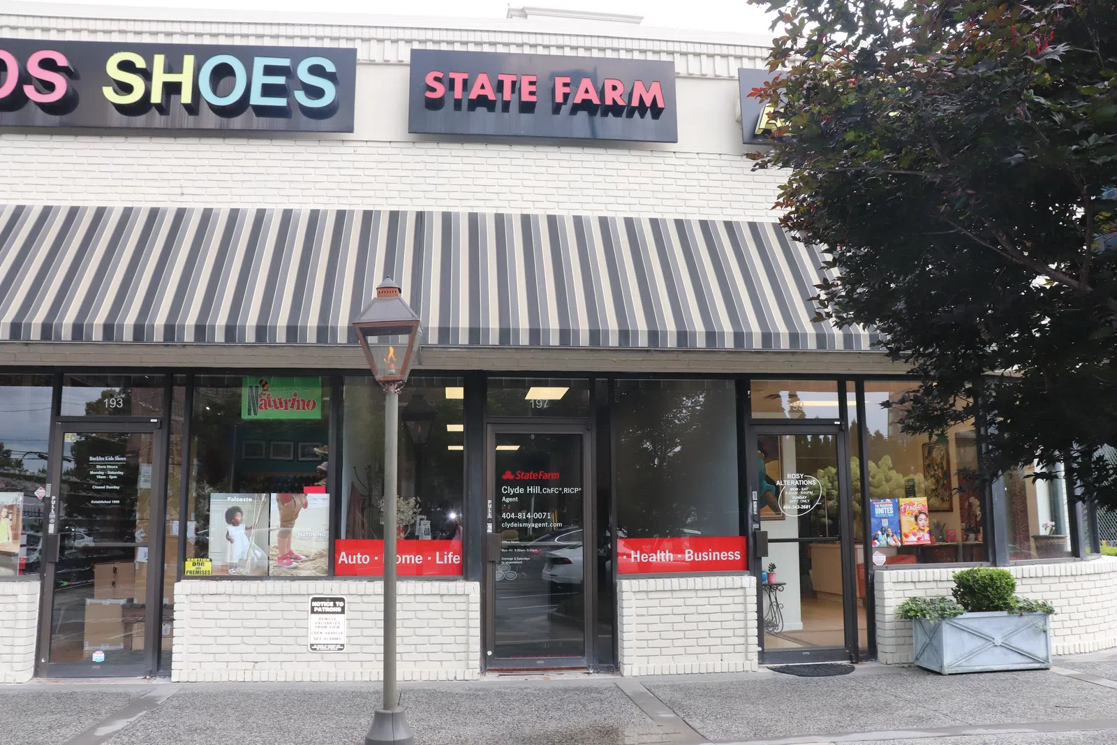 Strip mall storefronts featuring State Farm insurance office and Kids Shoes shop under a striped awning with street lamp.