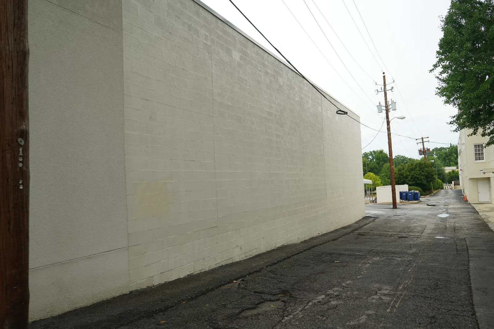 Empty urban alley beside a plain gray wall with cracked asphalt and power poles under overcast sky