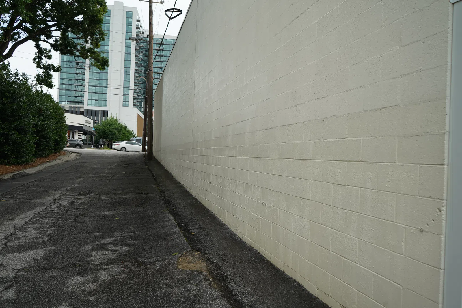 Empty urban alley with cracked pavement, beige brick wall, trees, and distant modern buildings under cloudy sky