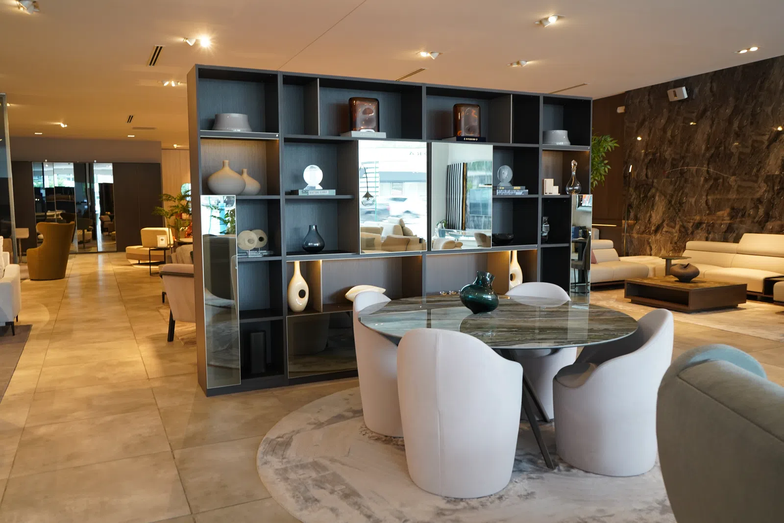 Modern living room with marble dining table, white chairs, open shelving, and neutral sofas in warm lighting
