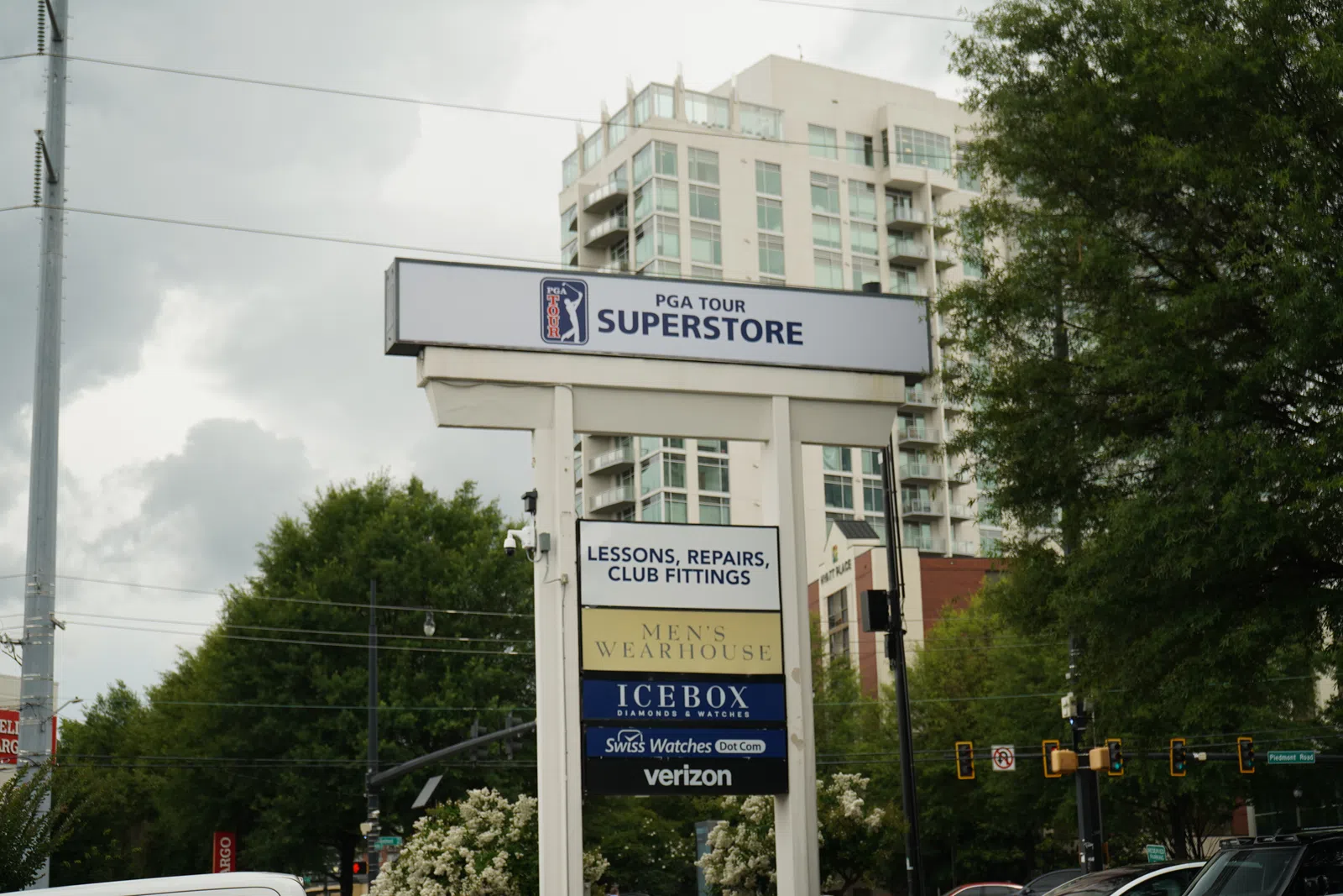 Outdoor multi-business sign for PGA Tour Superstore, Men's Wearhouse, Icebox, Verizon with city buildings and trees.