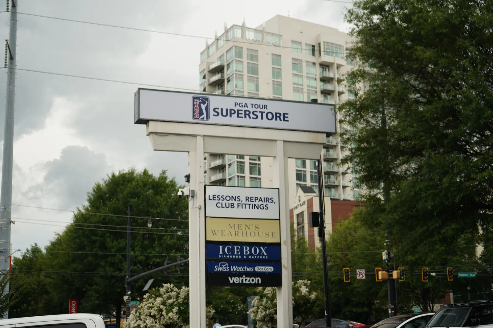 PGA Tour Superstore sign with multiple business ads against a cloudy sky and urban backdrop with trees and traffic lights.