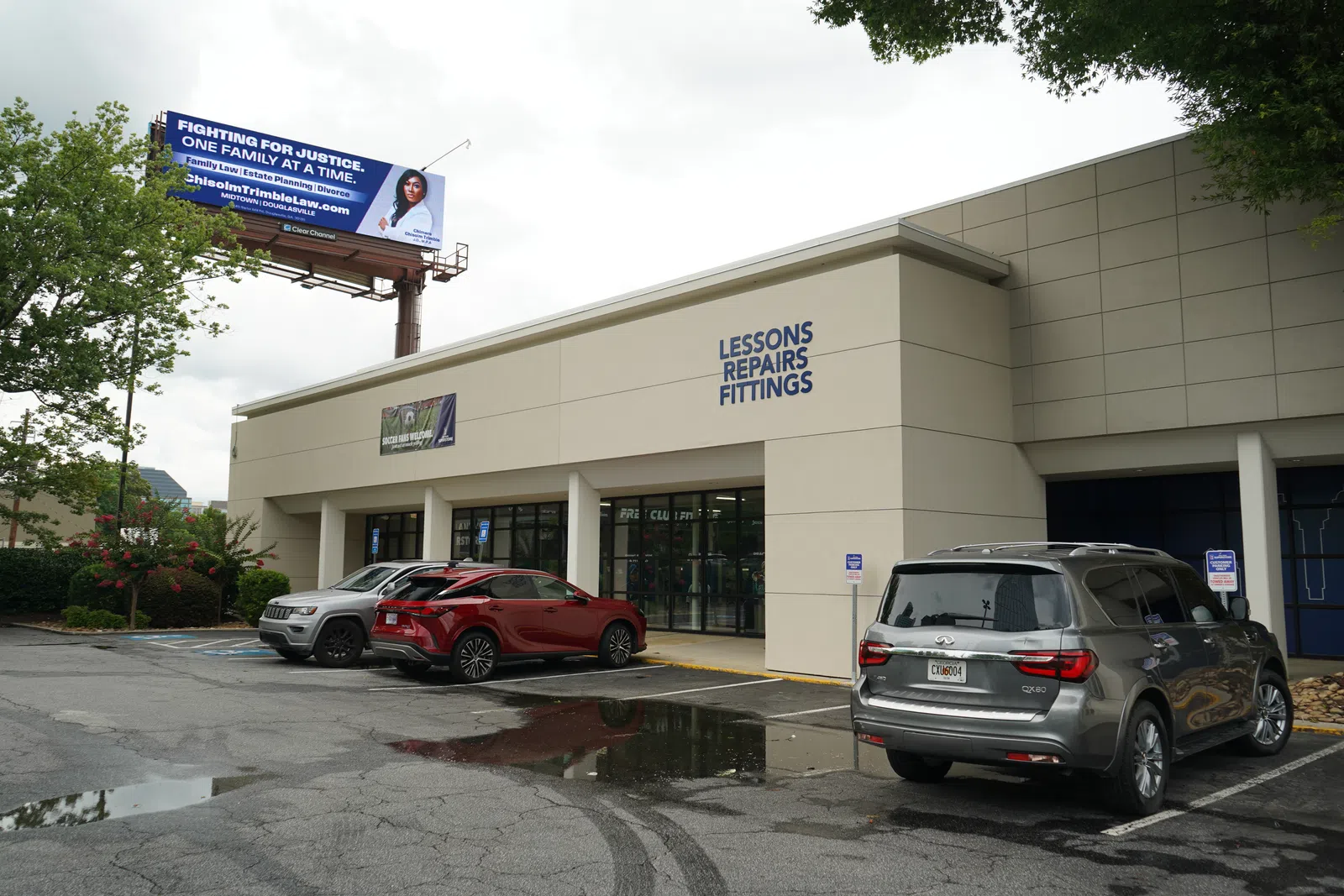 Storefront with signs for lessons, repairs, and fittings beside a parking lot with three cars and a justice billboard above.