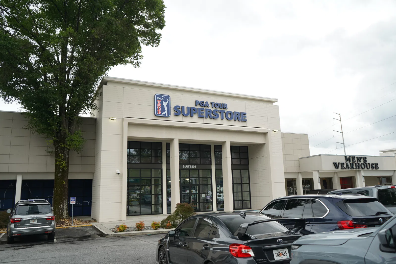 Front view of PGA Tour Superstore with parked cars and adjacent Men’s Wearhouse store in daylight