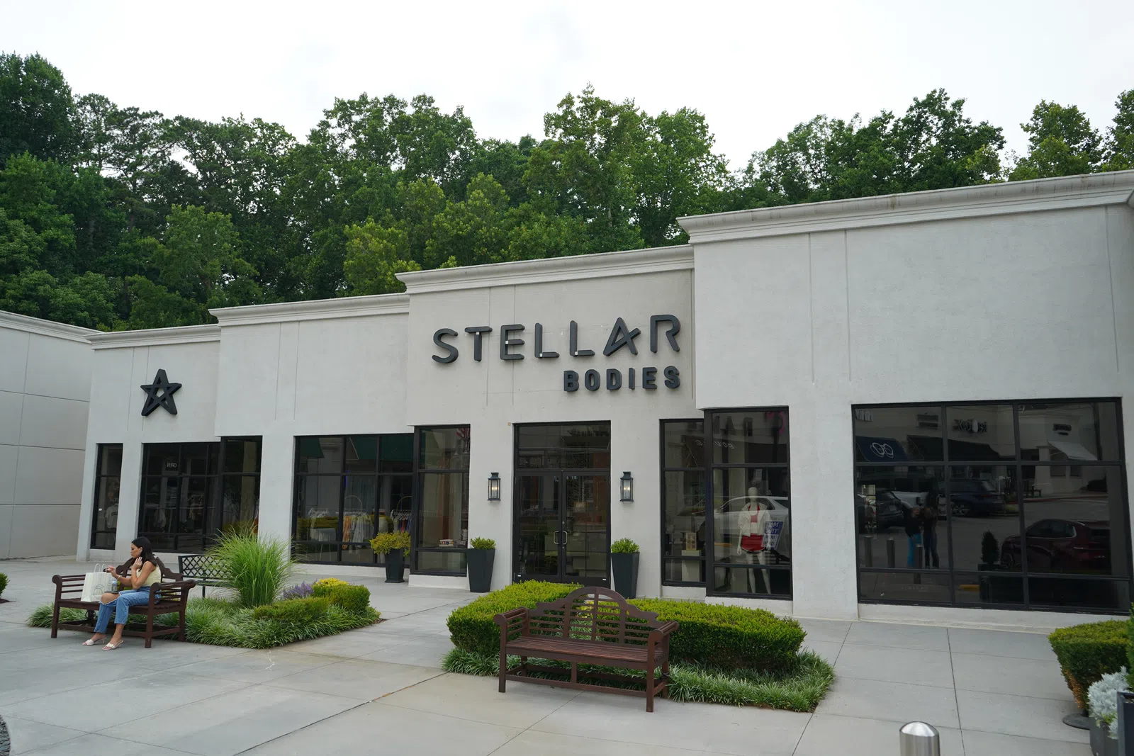 Exterior of Stellar Bodies store with large windows, entrances, benches, and greenery on a bright day.