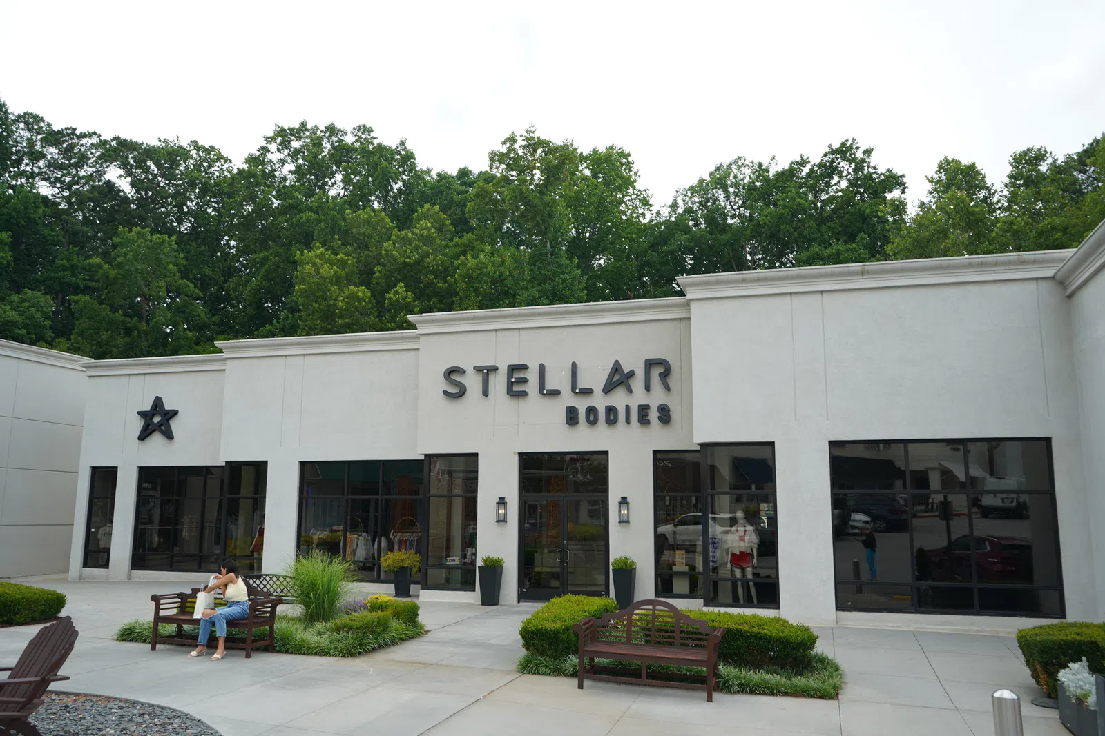 Stellar Bodies retail storefront with benches, potted plants, large glass windows, and green trees in the background.