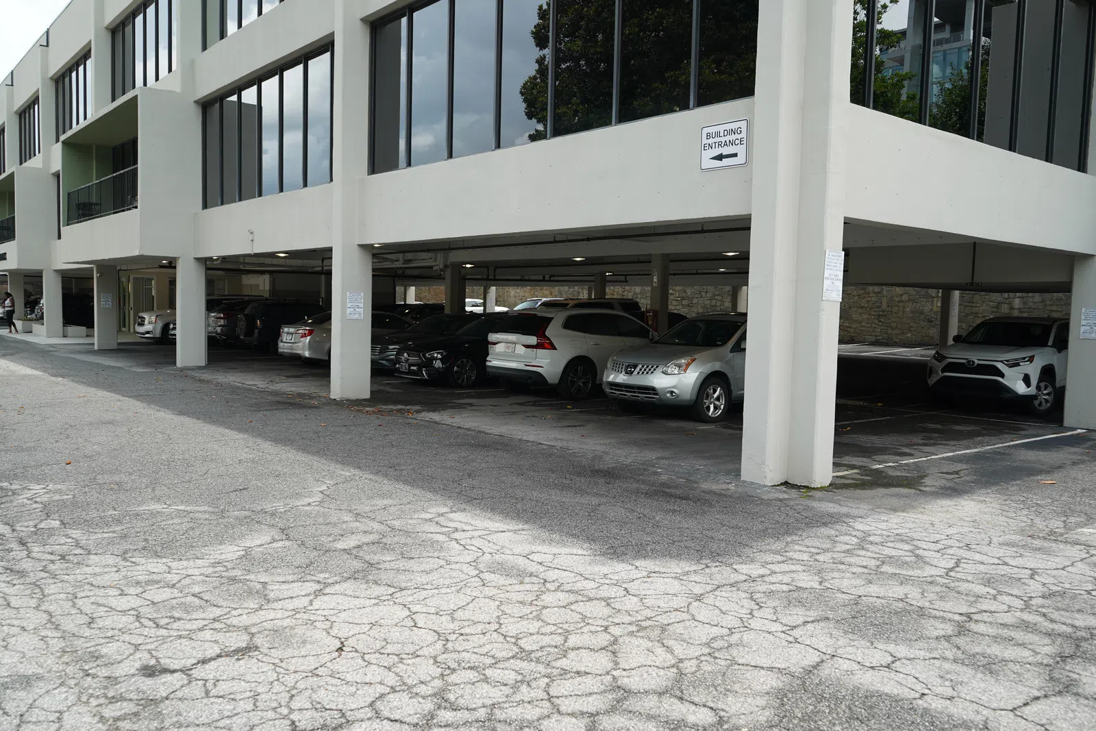 Cars parked under a modern office building with large windows and a cracked asphalt driveway.