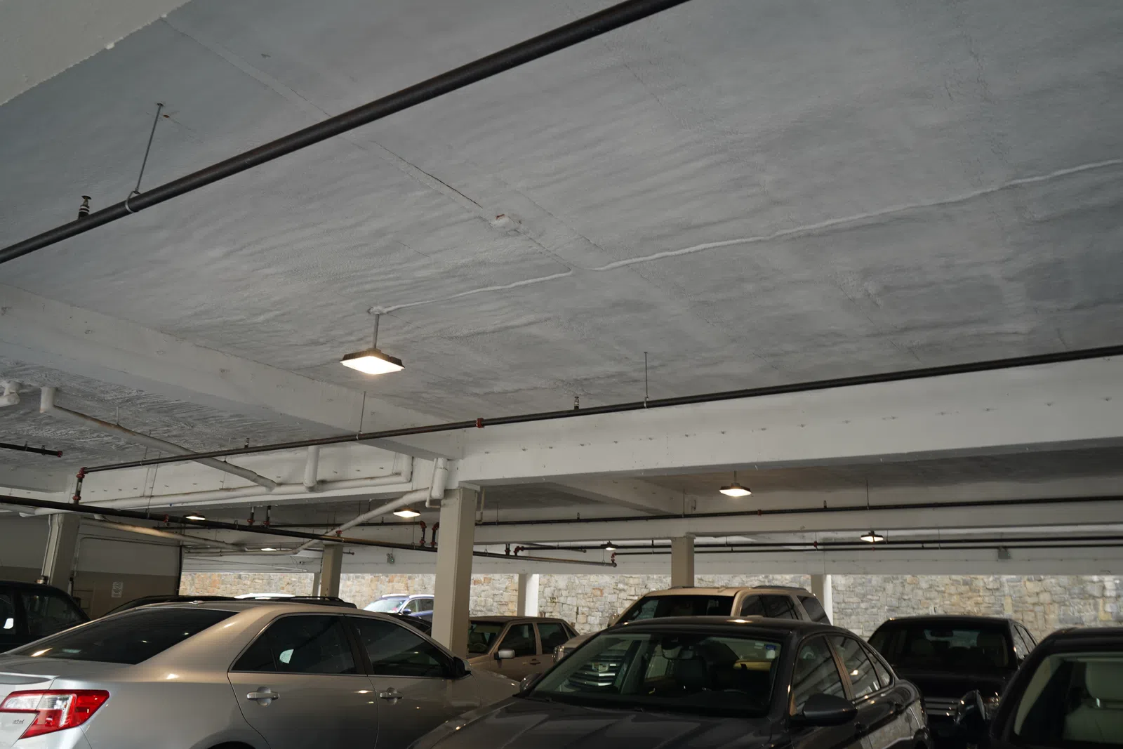Indoor parking lot with multiple cars parked under a concrete ceiling with overhead lights and pipes.