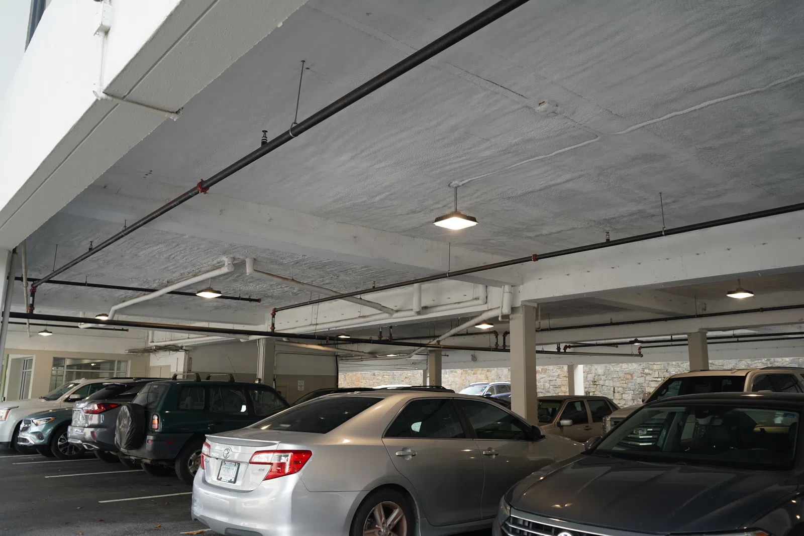 Indoor parking garage with multiple parked cars under a concrete ceiling with exposed pipes and lighting.