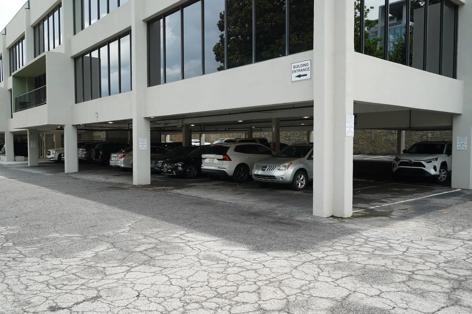 Covered parking lot under an office building with various cars parked and cracked asphalt pavement