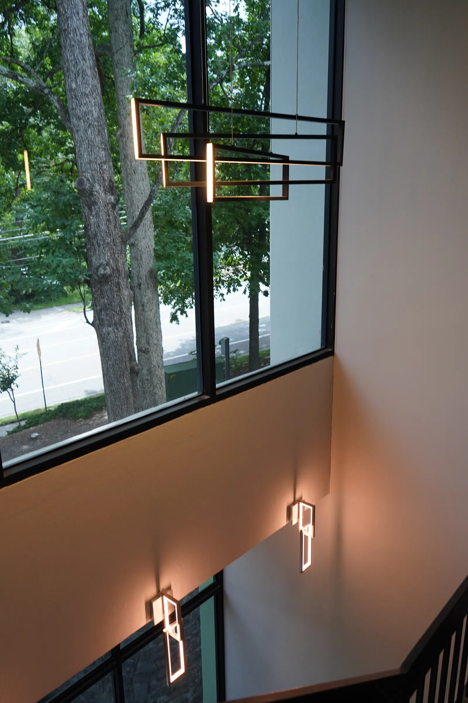 Modern geometric pendant lights and sconces illuminate a stairwell with large windows and green trees outside.
