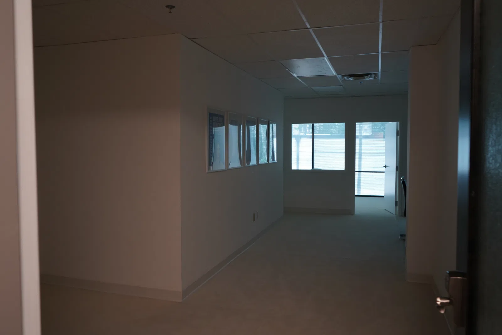 Dimly lit office hallway with framed pictures on the wall and large windows at the end with blinds.