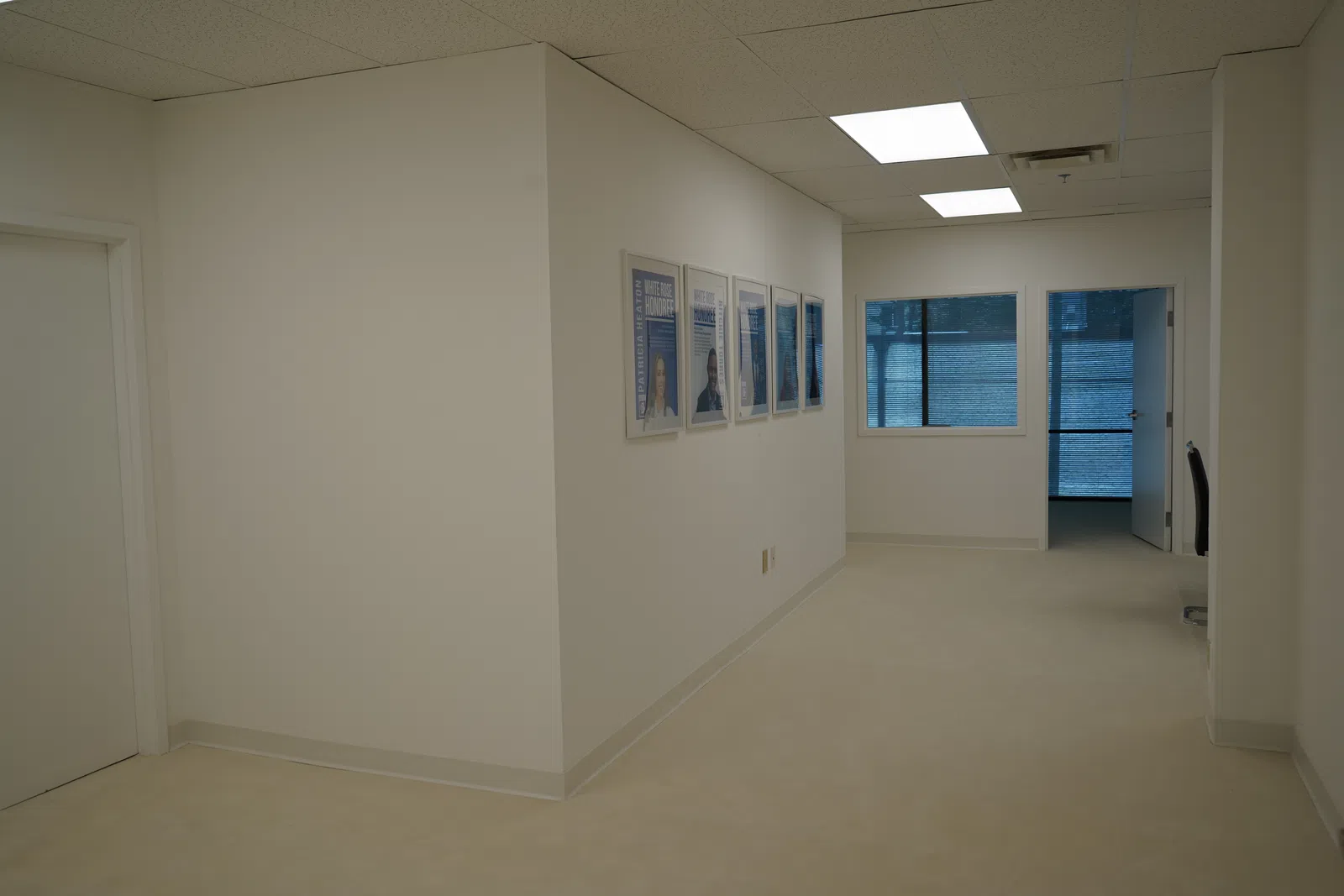 Bright office hallway with white walls, ceiling lights, framed pictures, and a window overlooking urban buildings.