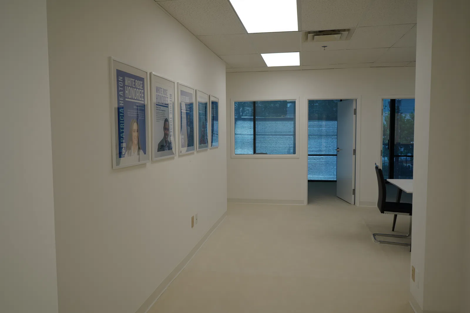 Bright office hallway with framed photos on wall and windows with blue blinds leading to rooms with chairs and tables.
