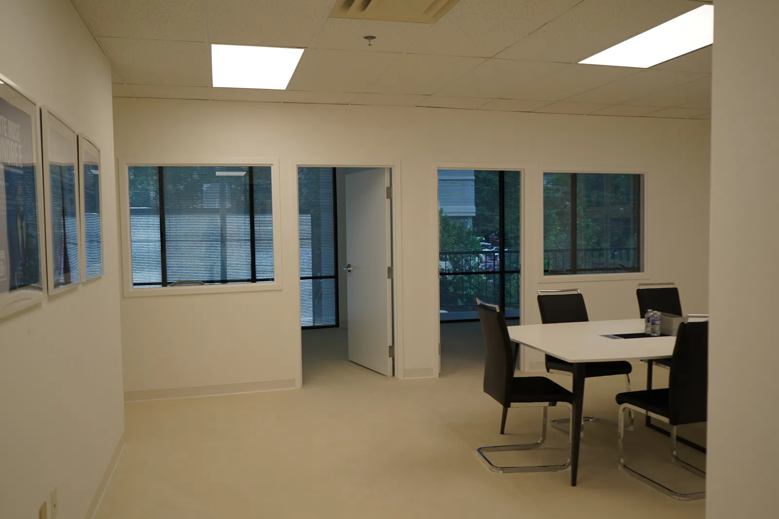 Bright, empty office room with beige carpet, white walls, table with black chairs, and windows showing outdoor greenery.