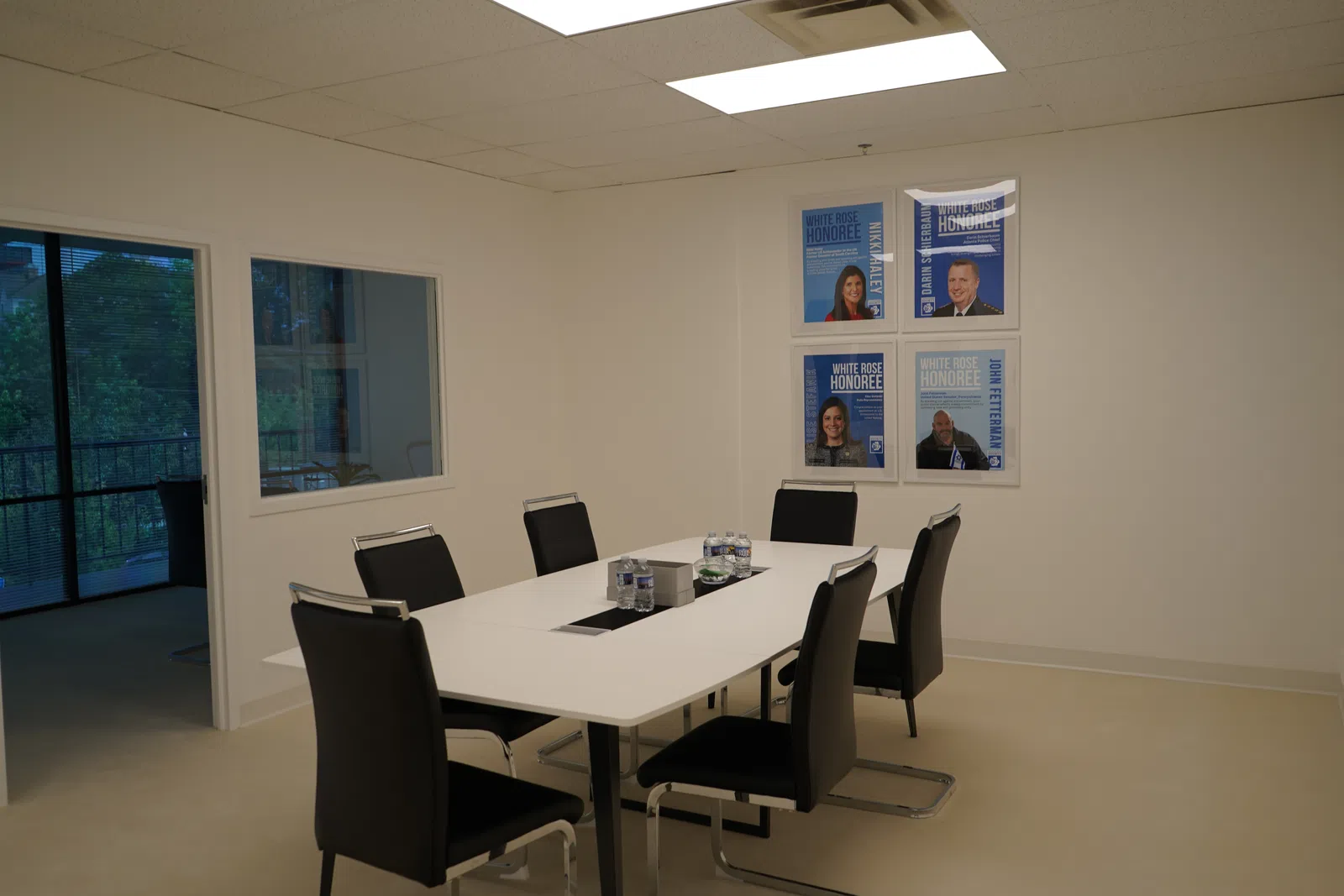 Modern conference room with white table, black chairs, water bottles, and framed honoree posters on the wall.