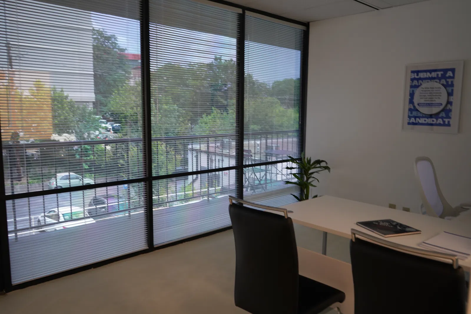 Modern office with large windows, blinds, black chairs, white desk, books, and a plant in natural light.